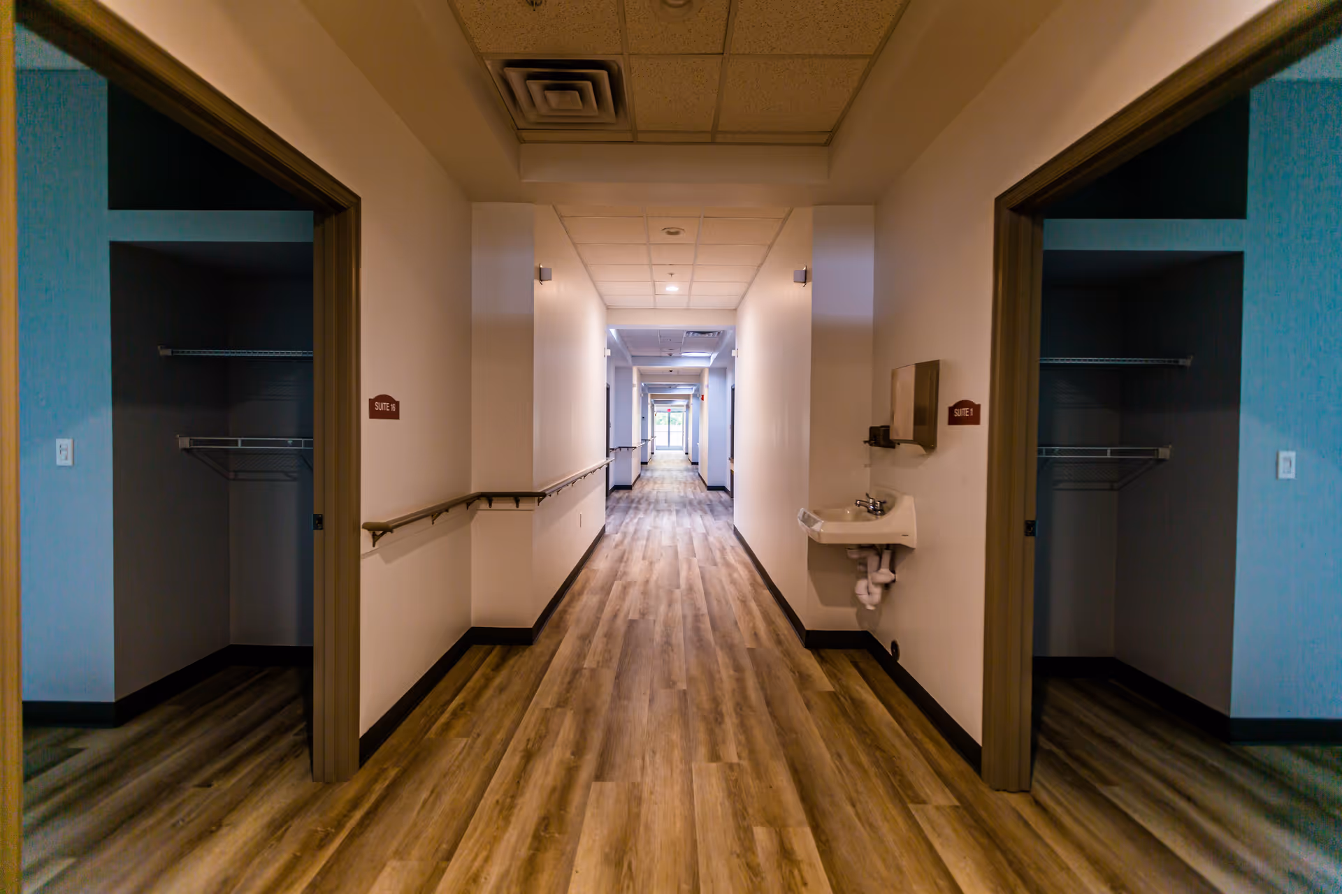 A long hallway in a senior living facility with wood-patterned flooring and white walls. There are open doorways on both sides leading to empty rooms with closet shelves. Handrails run along the walls of the hallway, and a small wall-mounted sink is visible on the right side near the front. The hallway is well-lit with ceiling lights and leads to a glass exit door at the far end.