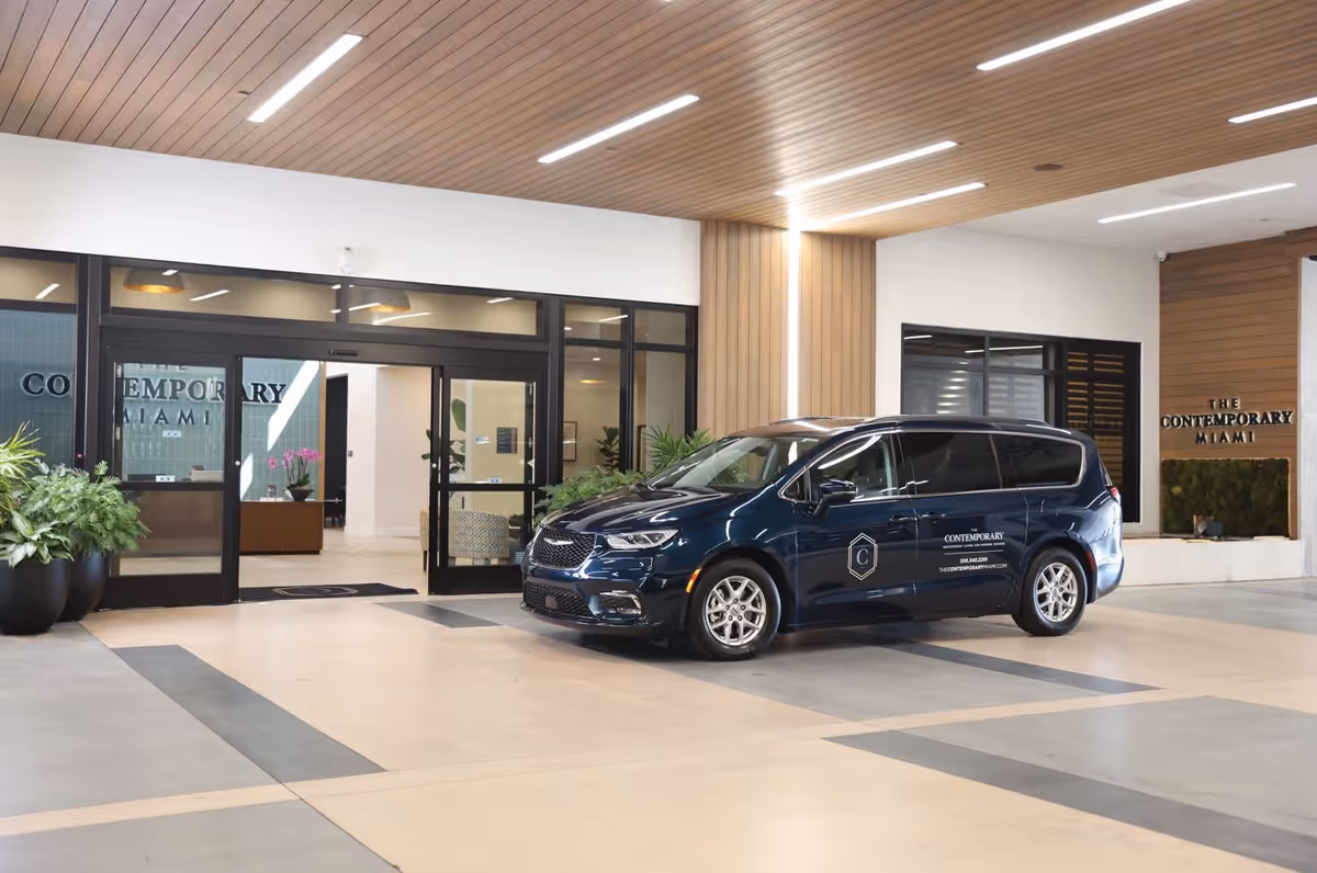 Entrance area of The Contemporary Miami facility showing a modern covered drop-off zone with a dark blue van parked in front. The van has branding for The Contemporary Miami on its side. The entrance features large glass doors and windows, potted plants, and a wooden ceiling with recessed lighting.