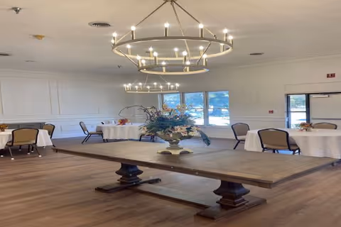 A spacious dining room with wooden flooring, round tables covered with white tablecloths, and chairs arranged around them. A large wooden rectangular table with a floral centerpiece is in the foreground. Modern circular chandeliers hang from the ceiling, and windows in the background allow natural light to enter the room.