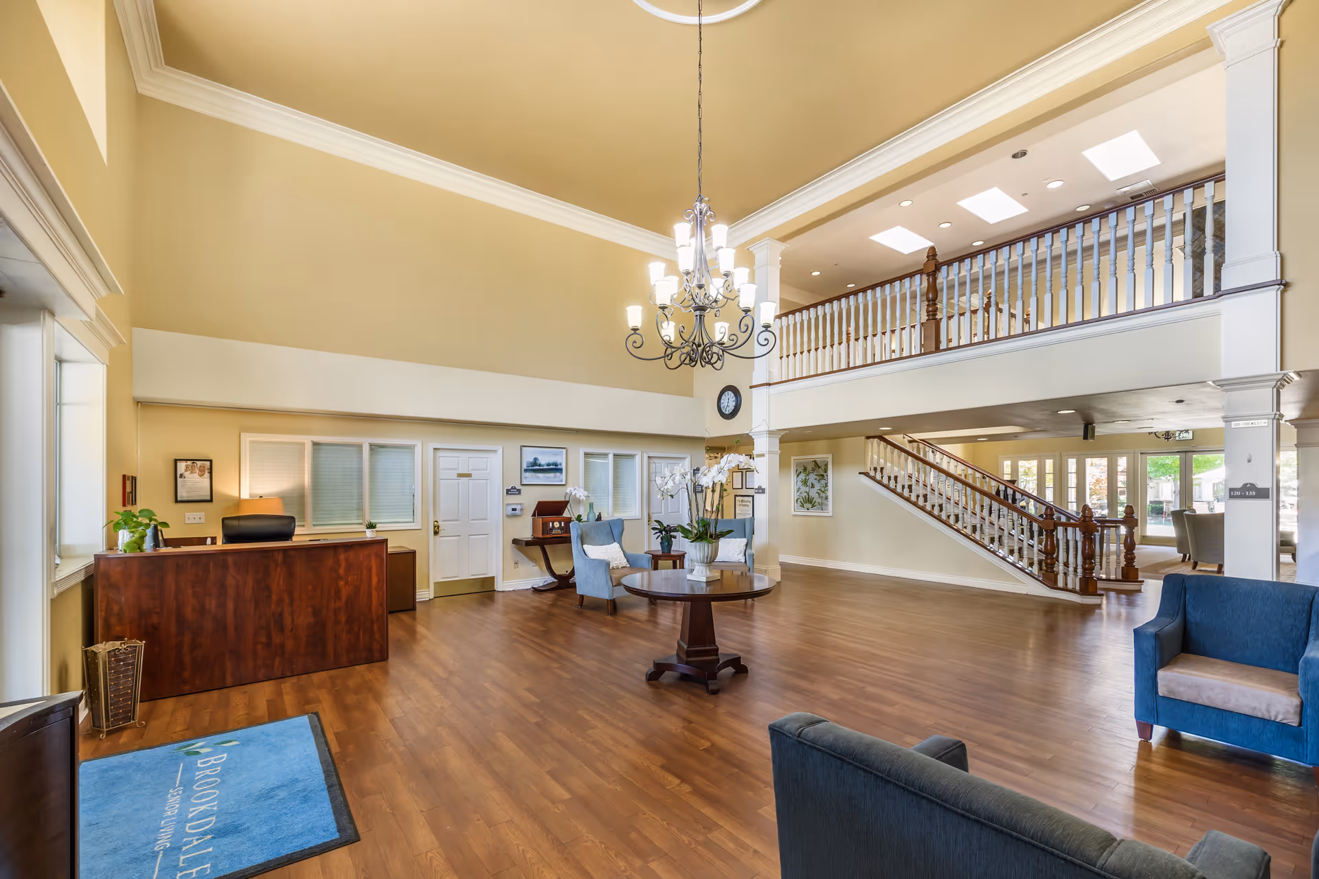 Spacious and well-lit senior living facility lobby with high ceilings, a chandelier, wooden flooring, a reception desk, seating area with blue armchairs, a round table with a flower arrangement, and a staircase leading to an upper level with white railings.