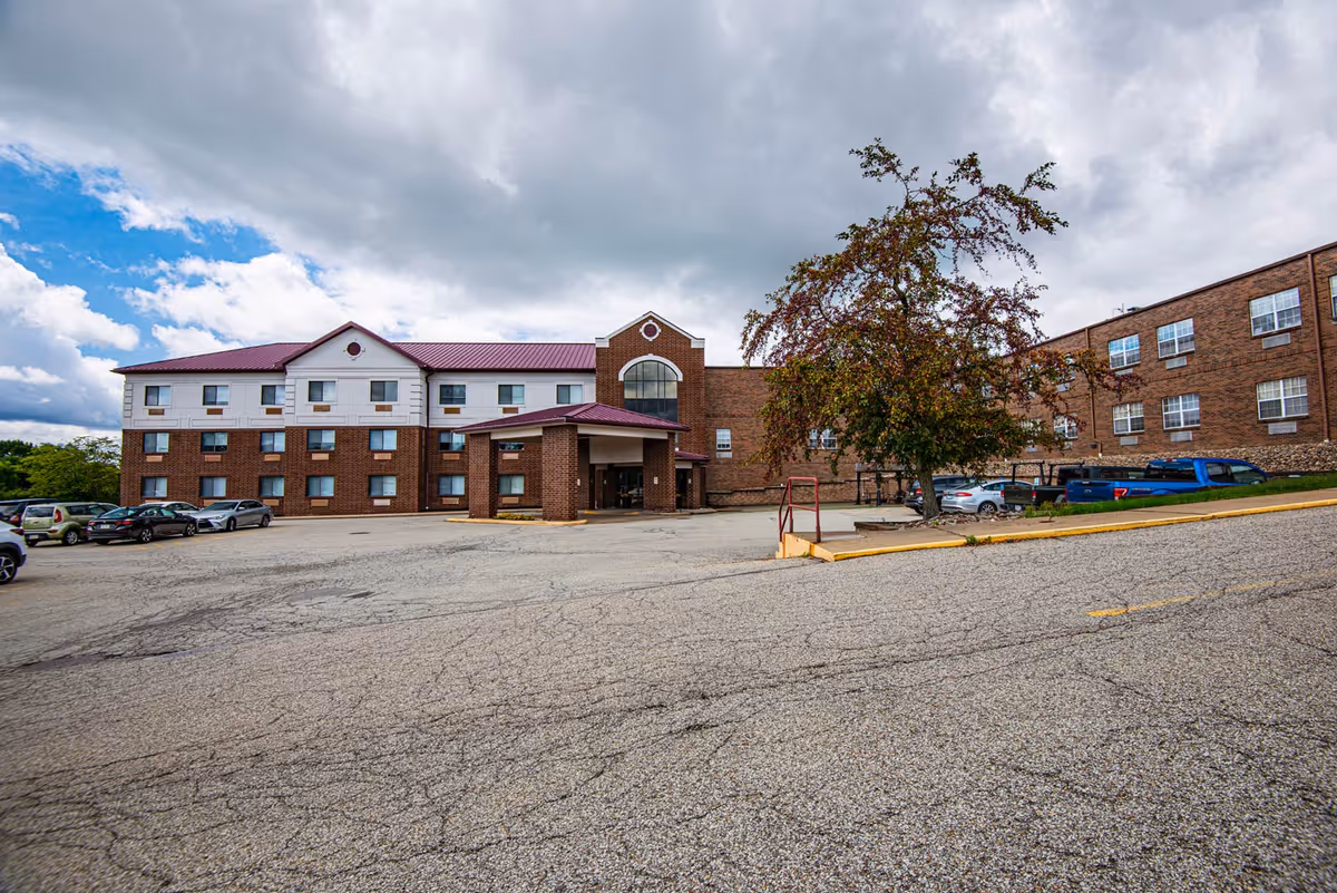 Exterior view of a three-story brick building with a maroon roof, several windows, and a covered entrance. There is a parking lot in front with several parked cars and a tree near the entrance. The sky is partly cloudy.