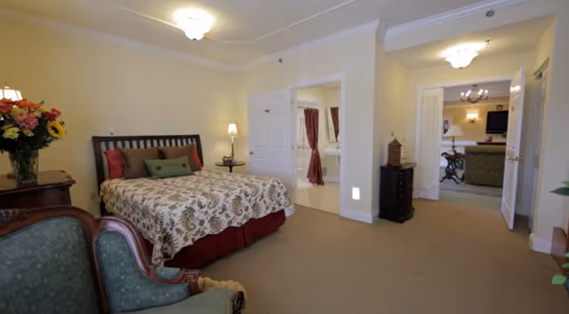 Spacious assisted living bedroom with a bed, armchair, dresser and open doorways to a bathroom and adjoining sitting area.