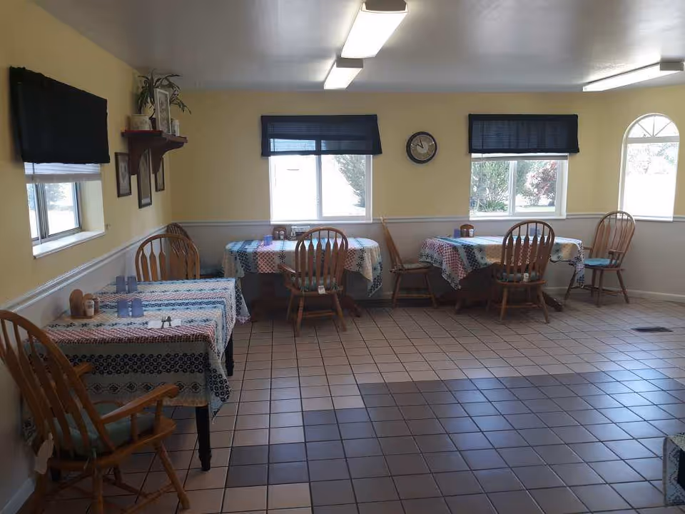 A dining room with several wooden tables covered with patterned tablecloths and wooden chairs. The room has tiled floors, yellow walls with white wainscoting, three windows with dark blue valances, a wall clock, and a mounted TV on the left wall.