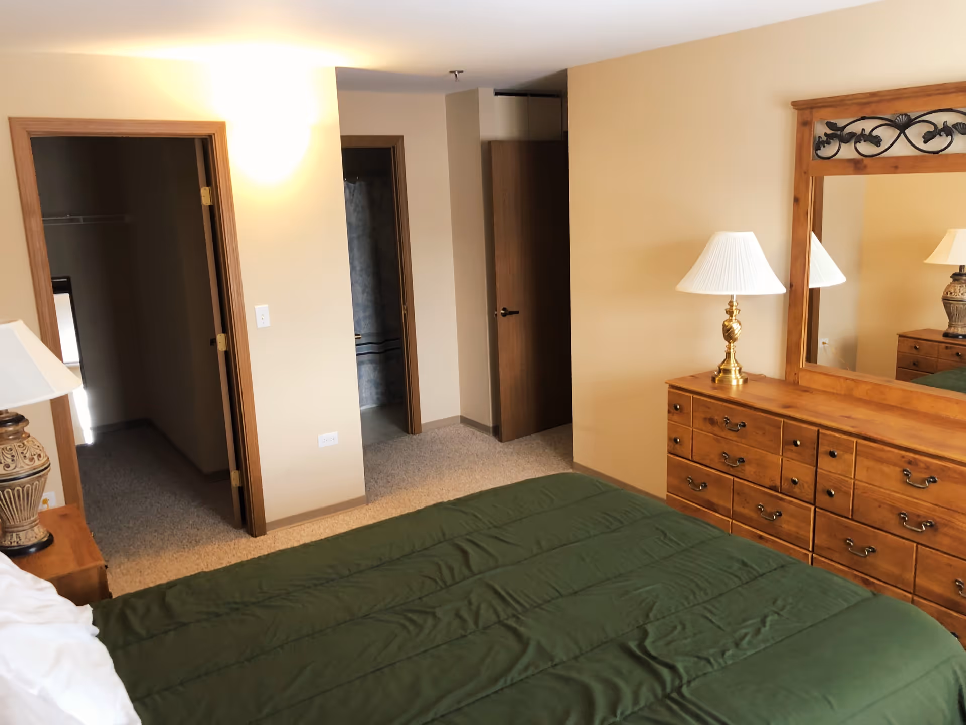A bedroom with a green bedspread on a bed, a wooden dresser with a large mirror, and a table lamp with a white shade. There are two open doorways visible, one leading to a closet and the other to a bathroom. The walls are painted beige and the floor is carpeted.