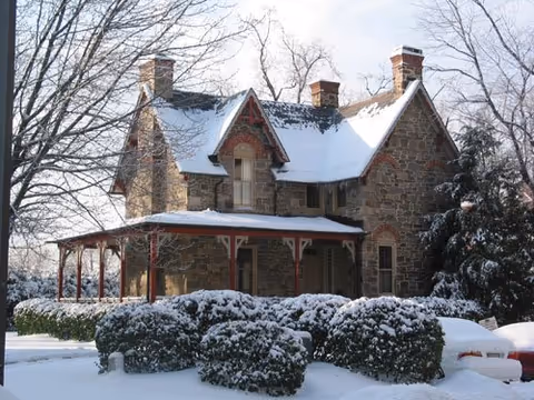 A two-story stone house with a steep roof covered in snow. The house has multiple chimneys and a wrap-around porch with wooden supports. Snow-covered bushes and trees surround the house, and the ground is blanketed with snow.