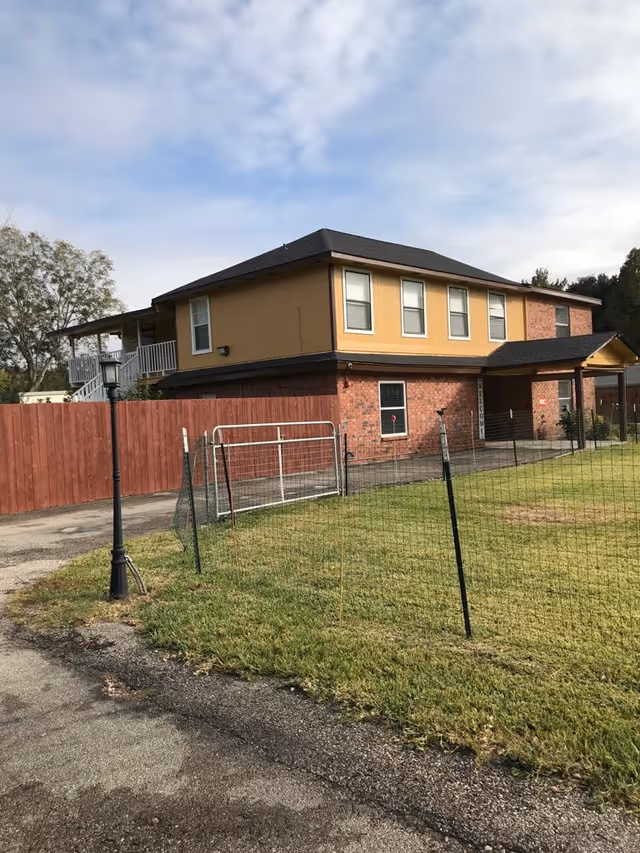 Two-story building with a brick lower level and yellow upper level, surrounded by a wooden fence and a wire fence on a grassy area under a partly cloudy sky.
