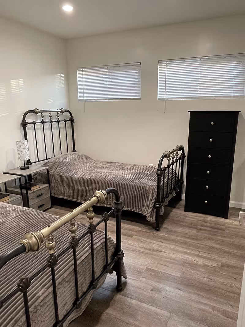 A bedroom with two single metal frame beds covered with gray striped blankets. Between the beds is a small nightstand with a lamp and some books. There is a tall black dresser against the wall. The room has light-colored walls, two horizontal windows with blinds, and wood-look flooring.