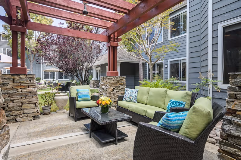 Outdoor seating area with a wicker sofa and two armchairs with green cushions and blue patterned pillows under a wooden pergola. A glass-top coffee table with a flower arrangement is in the center. Surrounding the area are stone pillars, plants, and trees, with a gray building in the background.