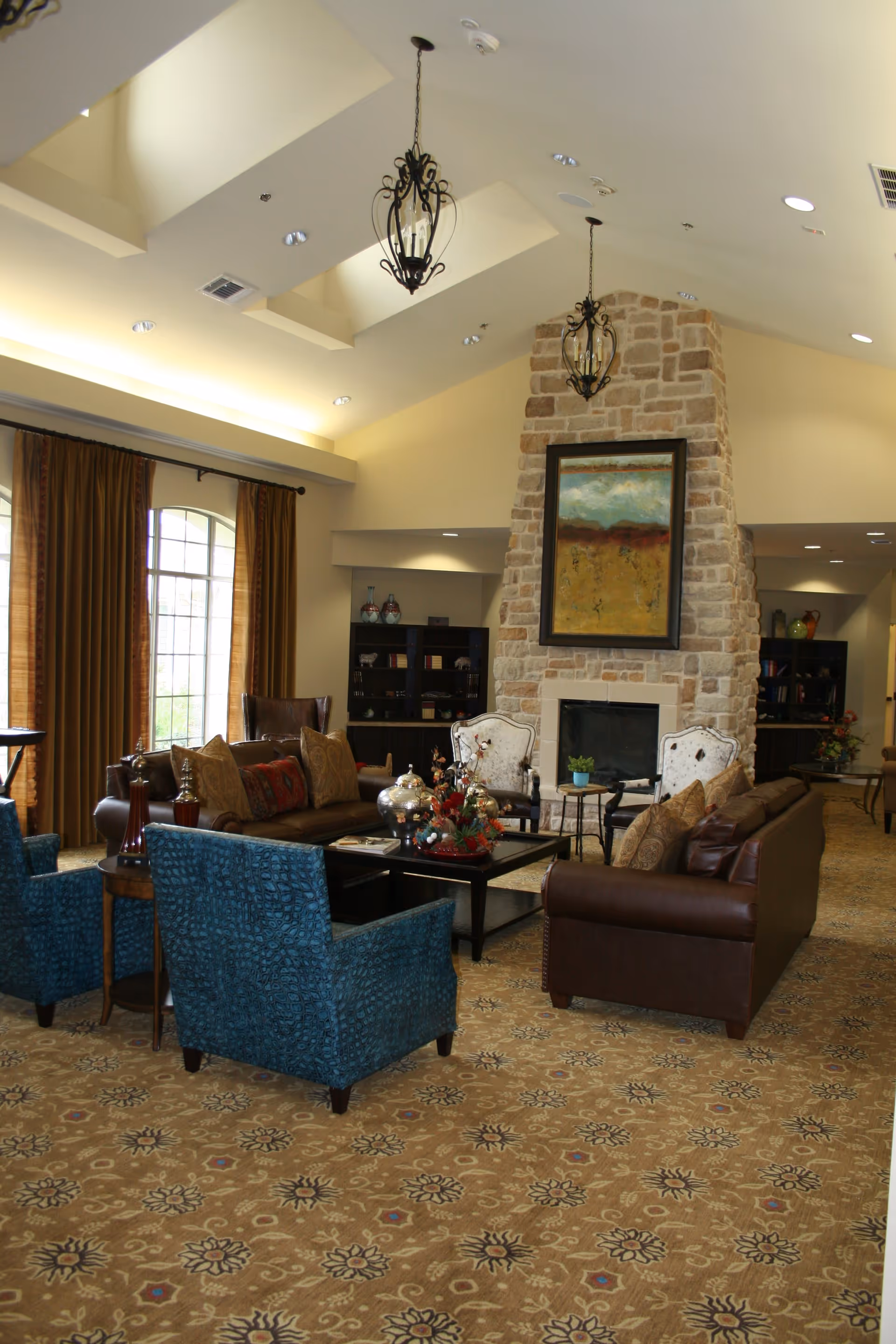 A spacious living room with high vaulted ceilings and two hanging decorative light fixtures. The room features a large stone fireplace with a colorful abstract painting above it. There are multiple seating options including brown leather sofas, blue upholstered armchairs, and two white cushioned chairs. A patterned carpet covers the floor, and large windows with brown curtains allow natural light to enter. Shelves with decorative items and books are visible in the background.