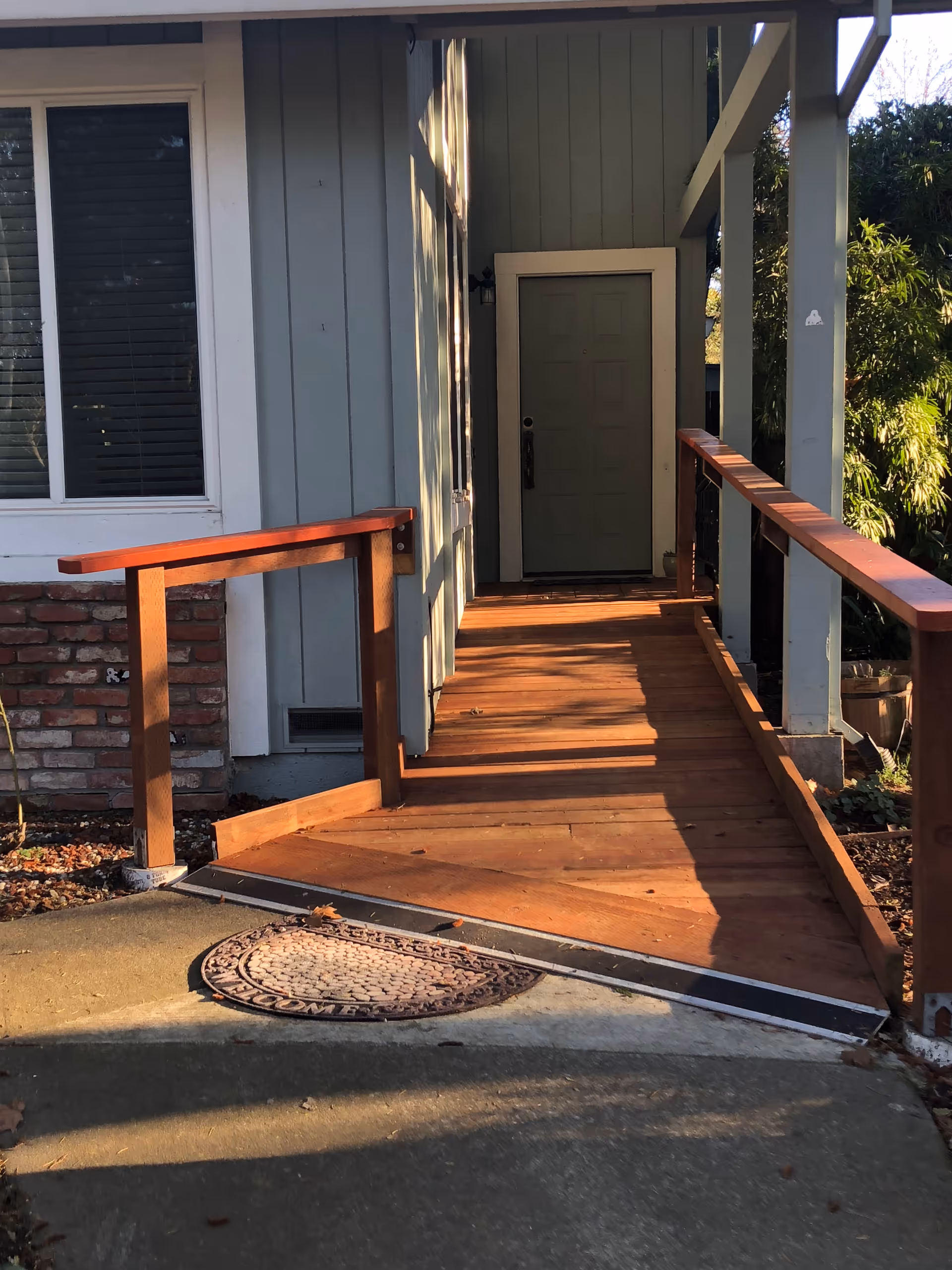 A wooden wheelchair ramp leading to a green front door of a house with light blue siding and a window with closed blinds on the left side. The ramp has wooden handrails on both sides and is surrounded by some greenery and a brick wall section.