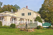 Exterior view of a two-story beige building with a white picket fence and green bushes in front. There is a green sign on the lawn that reads 'The Country Home Memory Care Specialist'. Trees with green foliage are visible in the background under a clear sky.