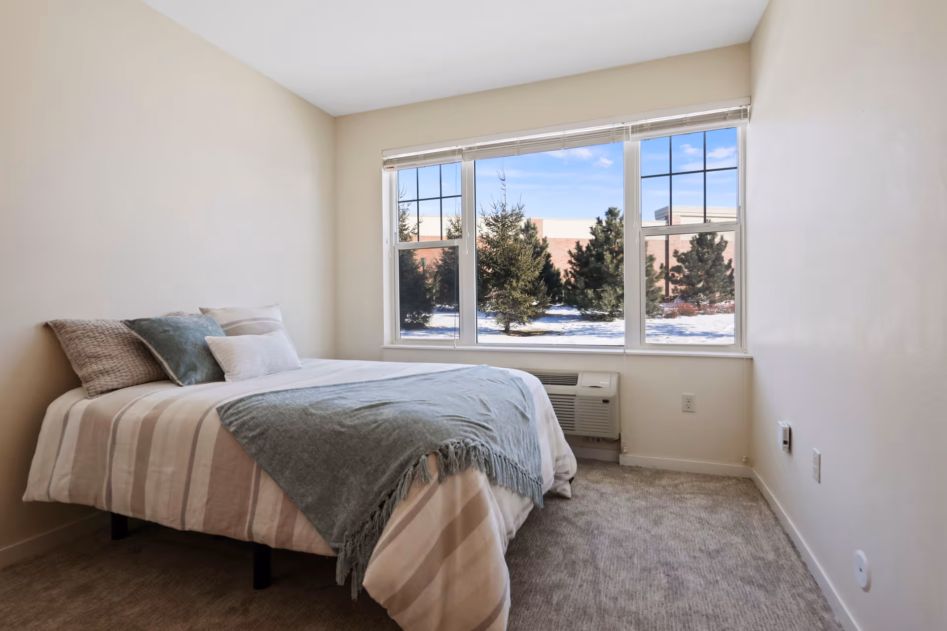 A bright bedroom with a neatly made bed beside a large window showing trees and snow outside.
