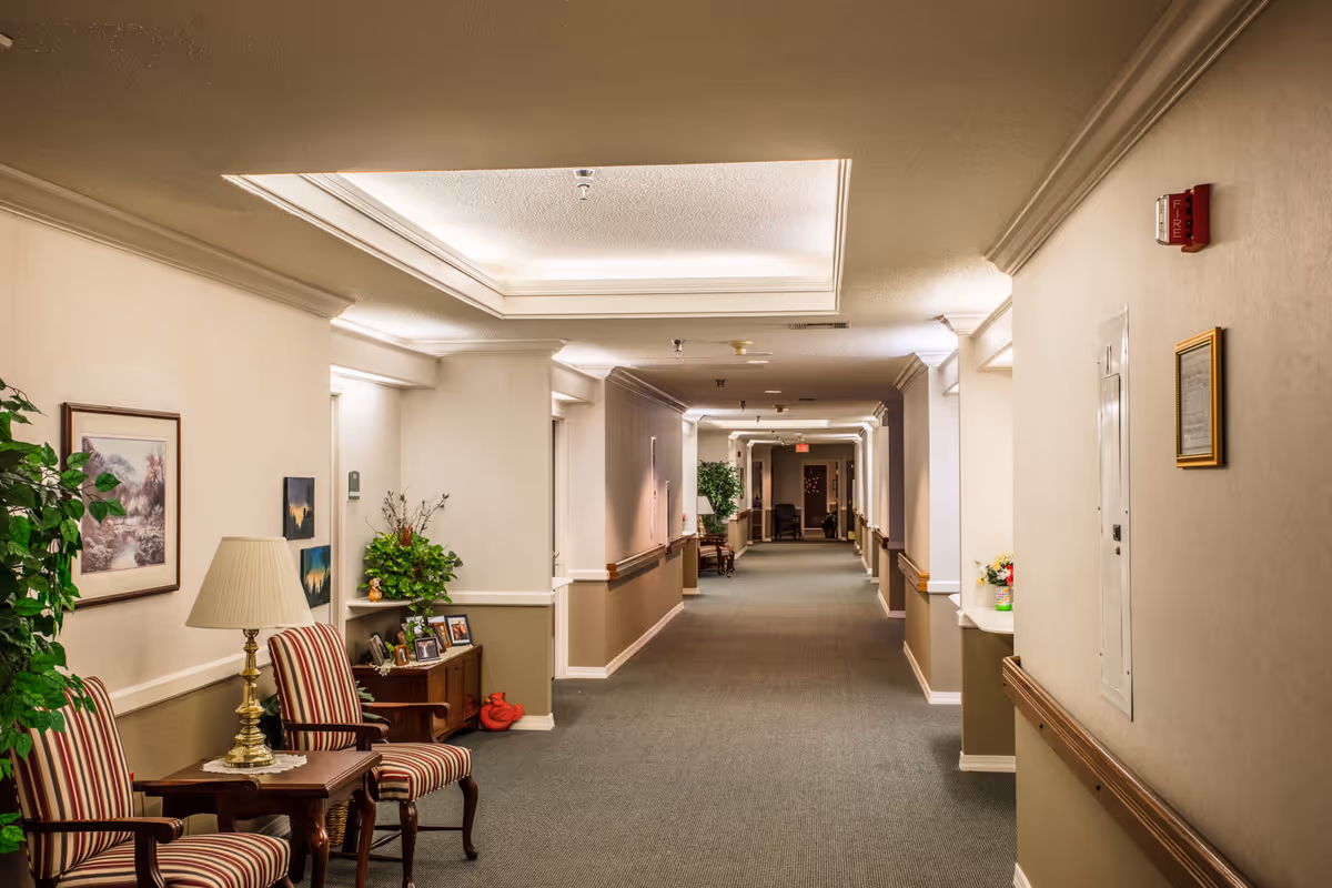 Long carpeted hallway in a senior living facility with seating areas, framed art, plants, and overhead lighting.