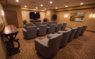 A cozy theater-style room with two rows of gray upholstered chairs facing a large flat-screen TV mounted on a beige wall. The room has warm lighting, a patterned carpet, a wooden side table, and a framed landscape painting on the wall.