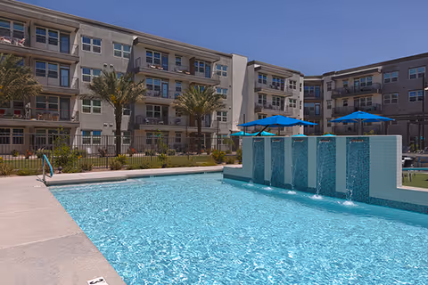 Outdoor swimming pool with clear blue water and water features in front of a multi-story residential building with balconies and palm trees under a clear blue sky.
