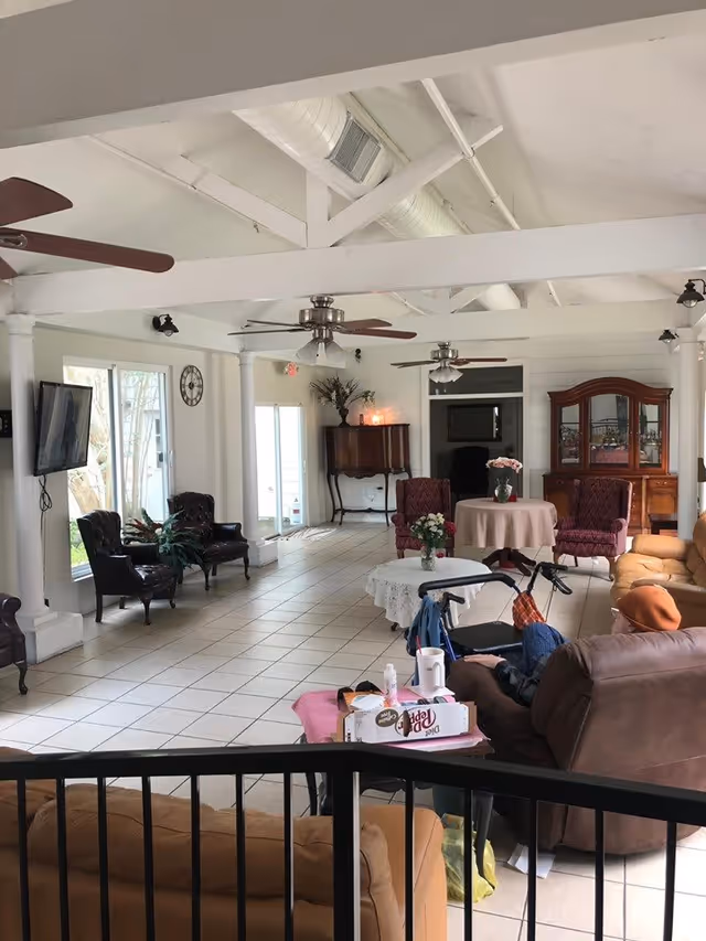 A spacious assisted living facility common area with white tiled floors and white walls. The room features multiple seating arrangements including leather armchairs, upholstered chairs, and sofas. There are ceiling fans and exposed white beams on the ceiling. A flat-screen TV is mounted on the wall near large windows letting in natural light. Tables with floral centerpieces and a wooden cabinet are also visible. A person is seated on a sofa with a walker nearby.