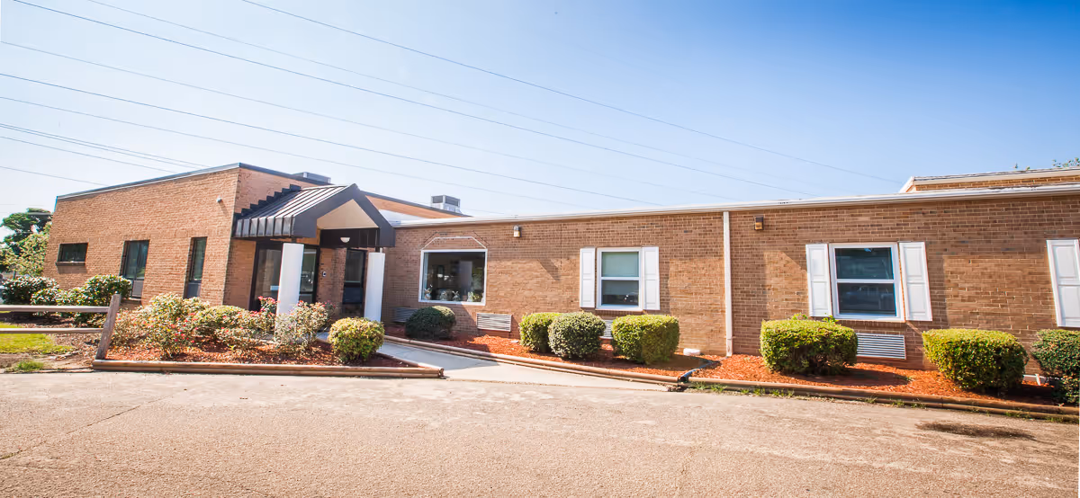 Exterior view of a single-story brick building with a covered entrance supported by white columns. There are several windows with white shutters and neatly trimmed bushes along the front. The sky is clear and blue.