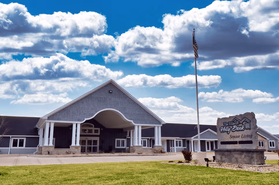 Exterior front view of Villas of Holly Brook Assisted Living & Memory Care building in Danville, IL, featuring a large covered entrance with white columns, a manicured lawn, an American flag on a flagpole, and a stone sign displaying the facility's name.