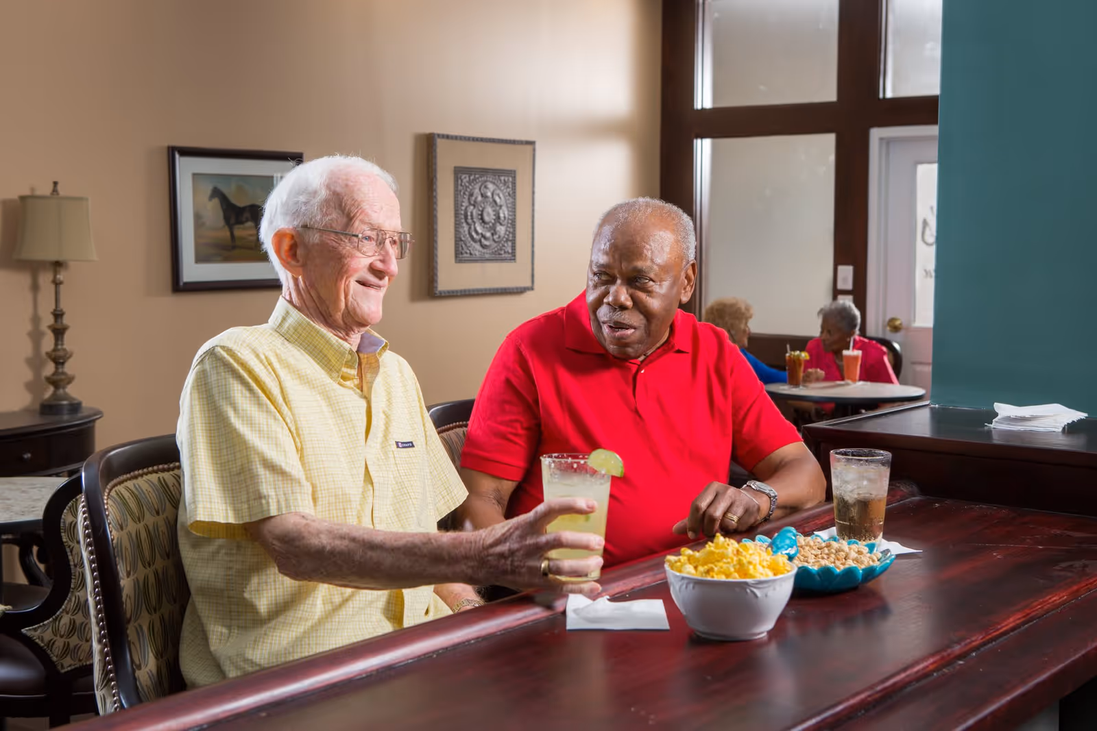 Two elderly men sitting at a wooden table in a common area, enjoying drinks and snacks. One man is wearing a yellow shirt and glasses, holding a drink with a lime garnish, while the other man in a red shirt looks on. In the background, two elderly women are seated at another table with drinks. The room has beige walls, framed artwork, and a lamp on a side table.
