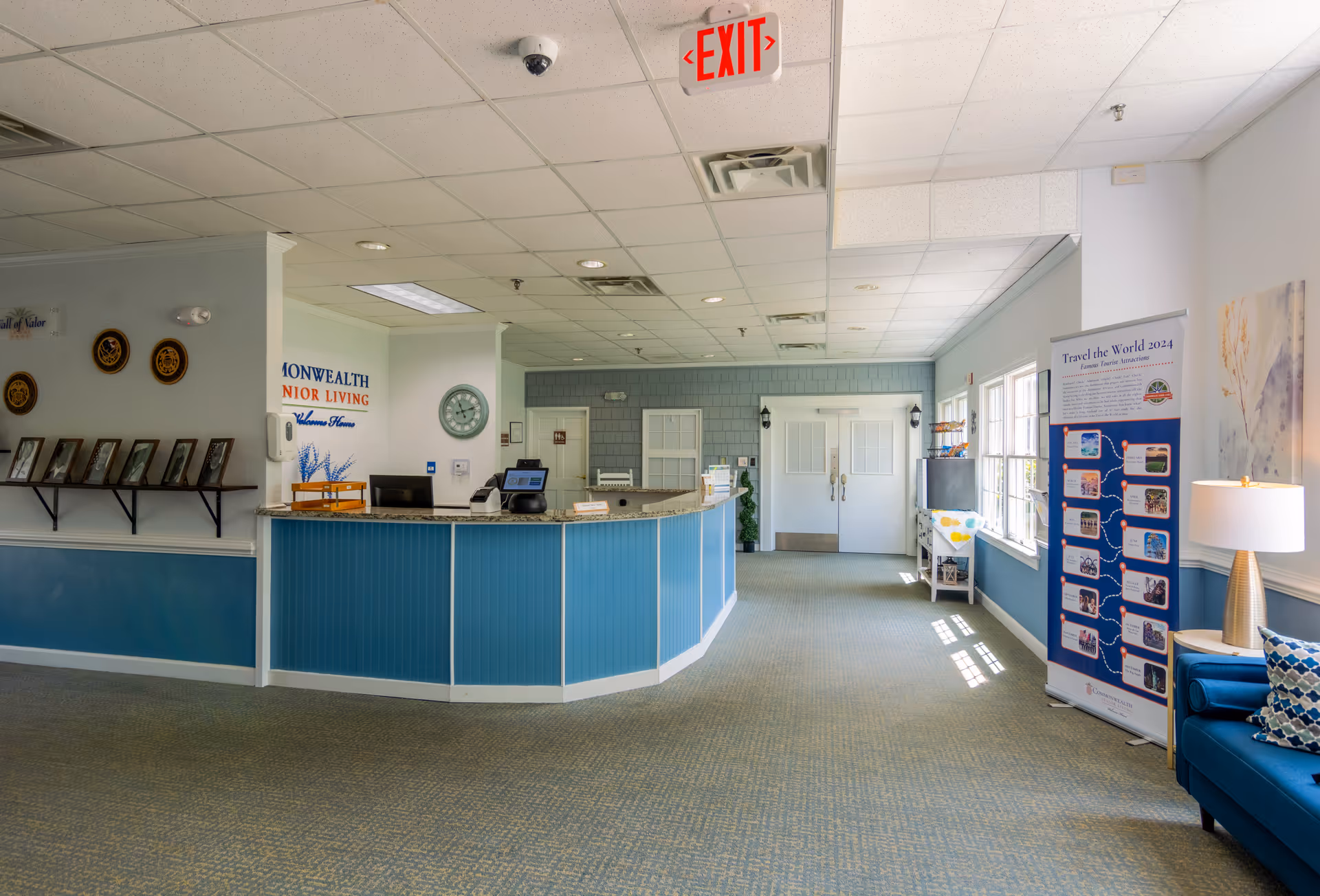 Reception area of a senior living facility with a blue front desk, a clock on the wall, a row of framed photos on a shelf, an exit sign on the ceiling, and a blue couch with patterned pillows near a lamp and a banner displaying Travel the World 2024 famous tourist attractions.