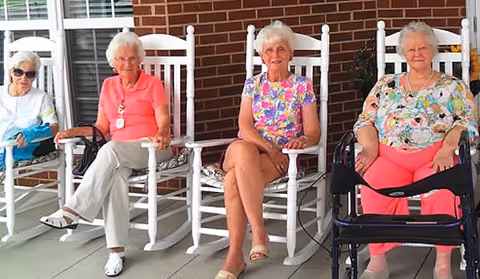 Four elderly women sitting on white rocking chairs on a covered porch with a brick wall background. One woman is using a walker, and they all appear relaxed and smiling.