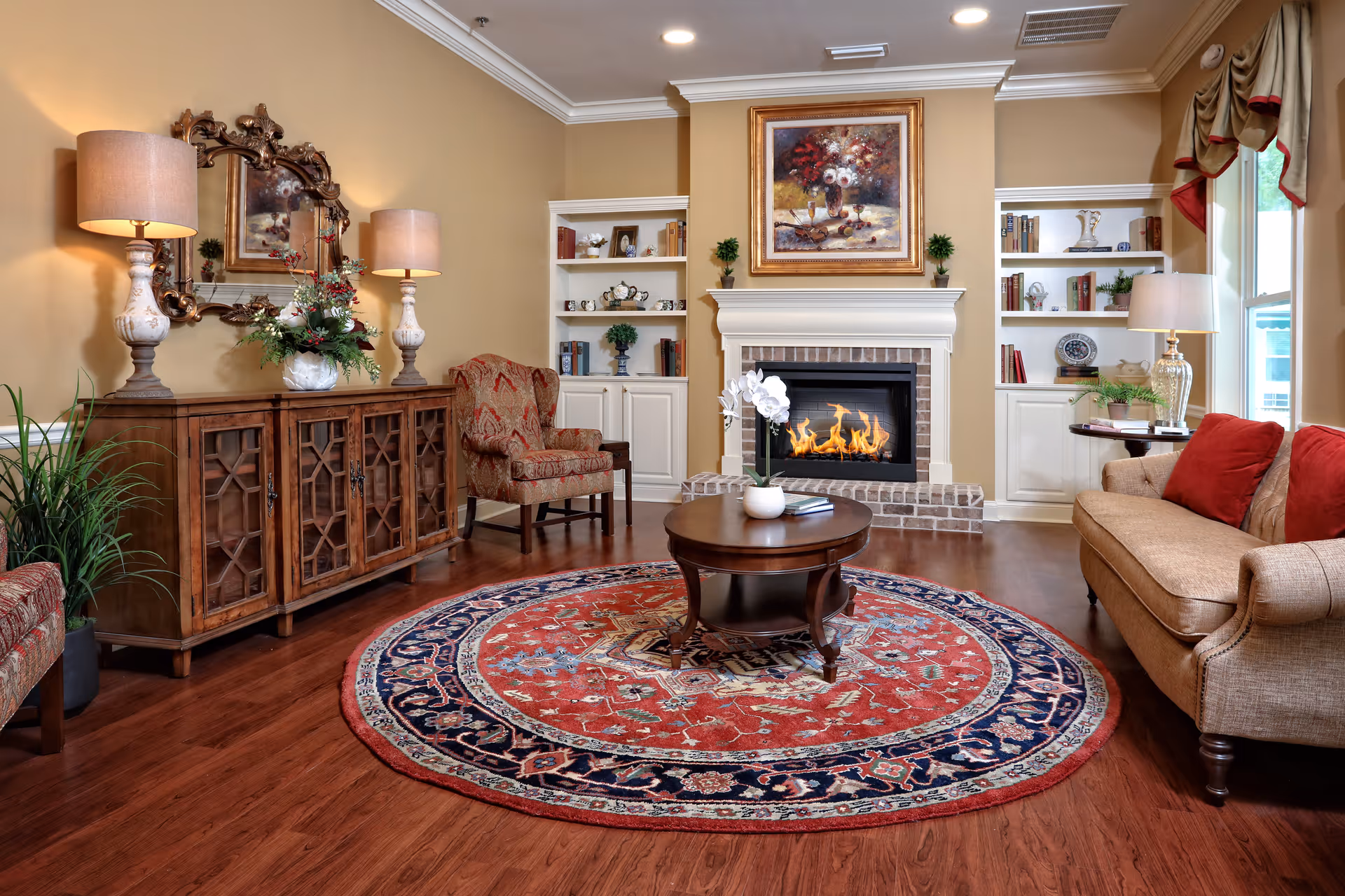 Cozy living room with a lit fireplace, round patterned rug, sofa, armchairs, and built-in bookshelves.