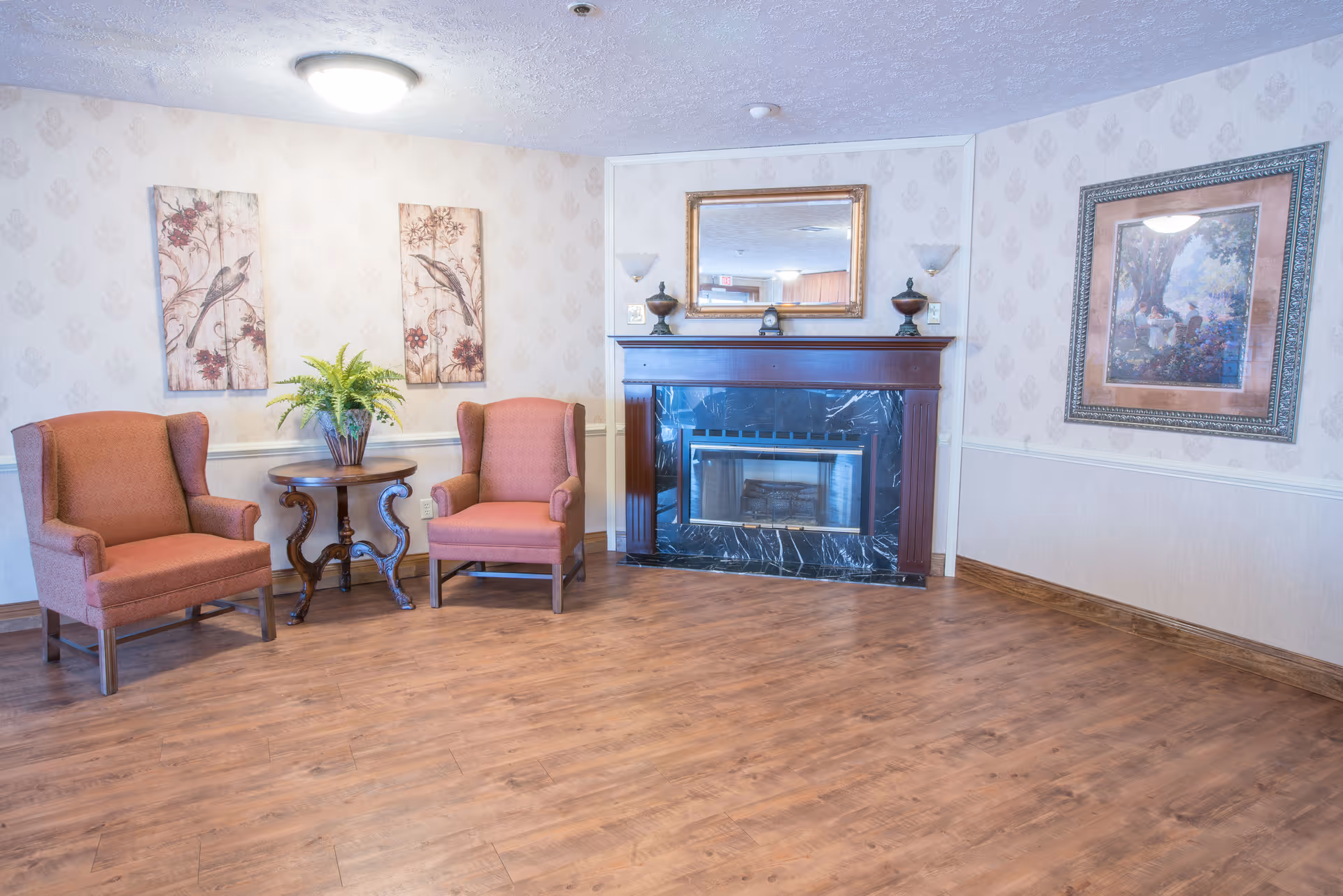 A cozy sitting area with two upholstered armchairs in a muted red color, separated by a small wooden table with a potted plant. The room features a fireplace with a dark marble surround and a wooden mantel, above which hangs a large mirror. The walls are decorated with patterned wallpaper and framed artwork. The floor is wood with a warm finish.
