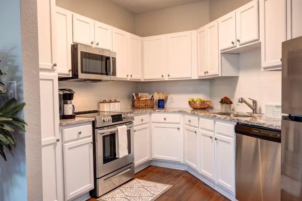 Modern kitchen with white cabinets, stainless steel appliances including a microwave, stove, dishwasher, and refrigerator. The countertop is granite with a bowl of green apples, a small plant, and kitchen utensils. There is a coffee maker on the counter and a patterned rug on the wooden floor.