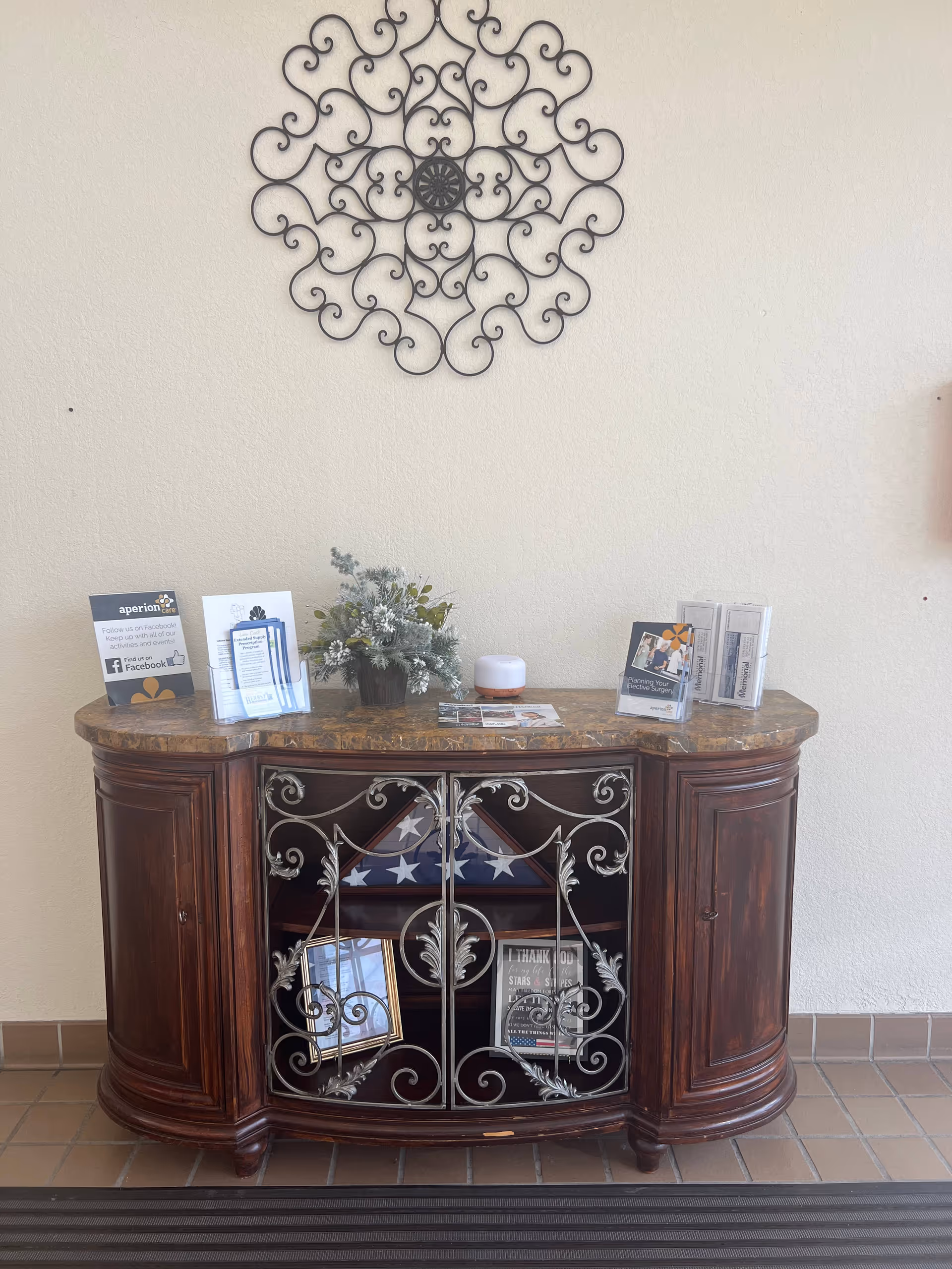 A decorative wooden cabinet with a marble top against a beige wall. On top of the cabinet are informational brochures, a small flower arrangement, and a diffuser. Above the cabinet is an ornate metal wall decoration. Inside the cabinet behind decorative metal doors is a folded American flag and framed pictures.