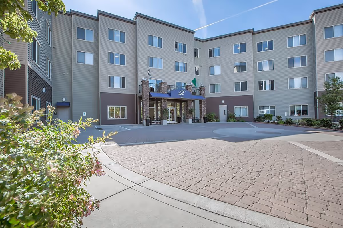 Exterior view of a multi-story senior living facility building with a paved driveway and entrance canopy. The building has multiple windows and a mix of beige and brown siding. There are some plants and shrubs near the entrance and along the driveway.