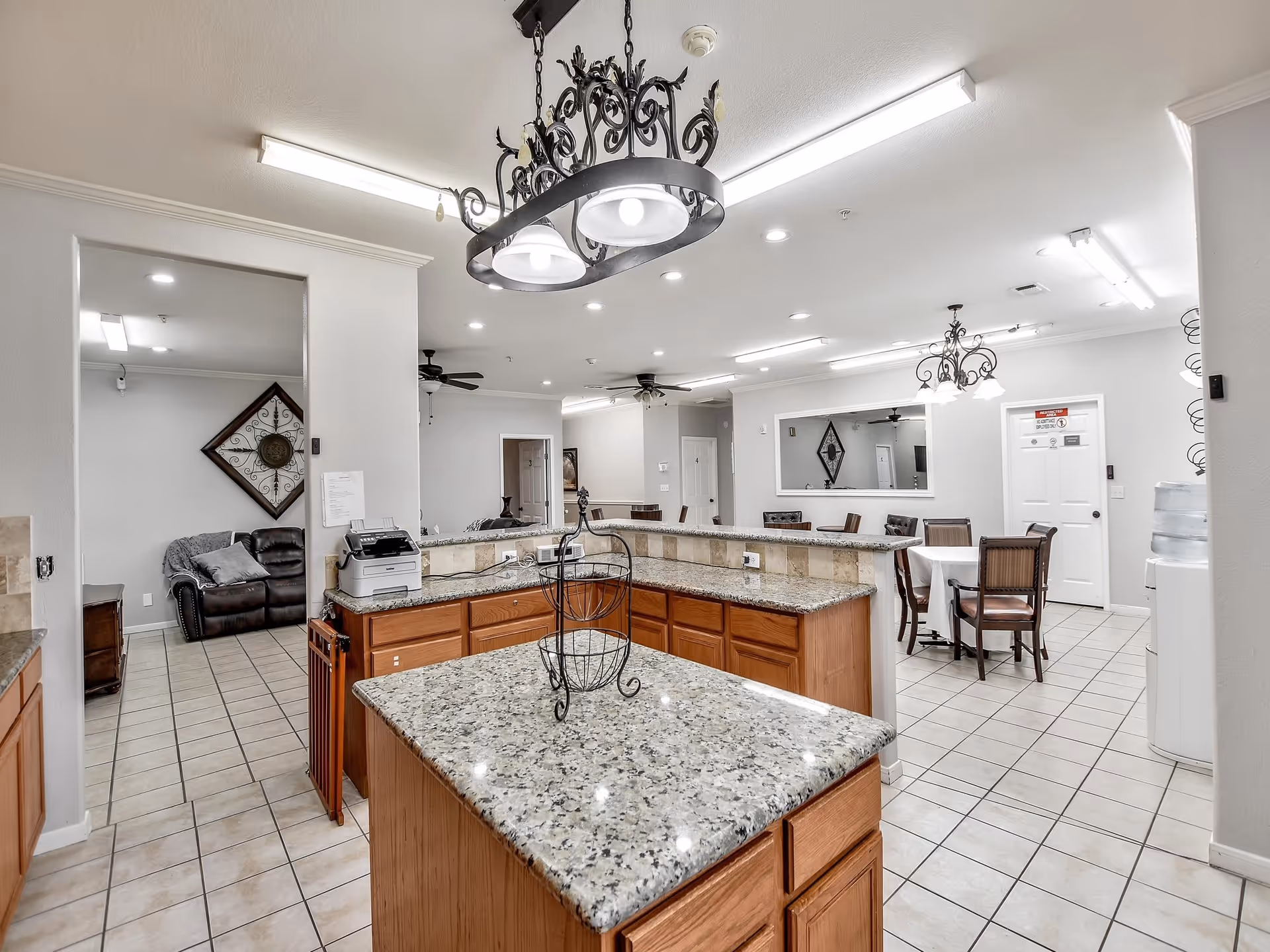 Interior view of a senior living facility showing a kitchen area with granite countertops and wooden cabinets. There is a decorative black metal chandelier hanging from the ceiling. In the background, there is a dining area with a table and chairs, a water cooler, and a doorway with a sign. To the left, there is a small sitting area with a leather couch and wall decoration.