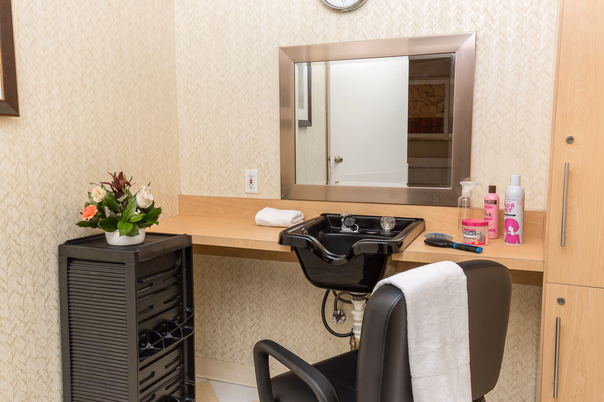 Interior view of a hair washing station in a convalescent hospital, featuring a black salon chair with a white towel draped over it, a black sink for hair washing, a large mirror with a silver frame, various hair care products on the counter, a small black storage unit with a flower arrangement on top, and light-colored patterned wallpaper.