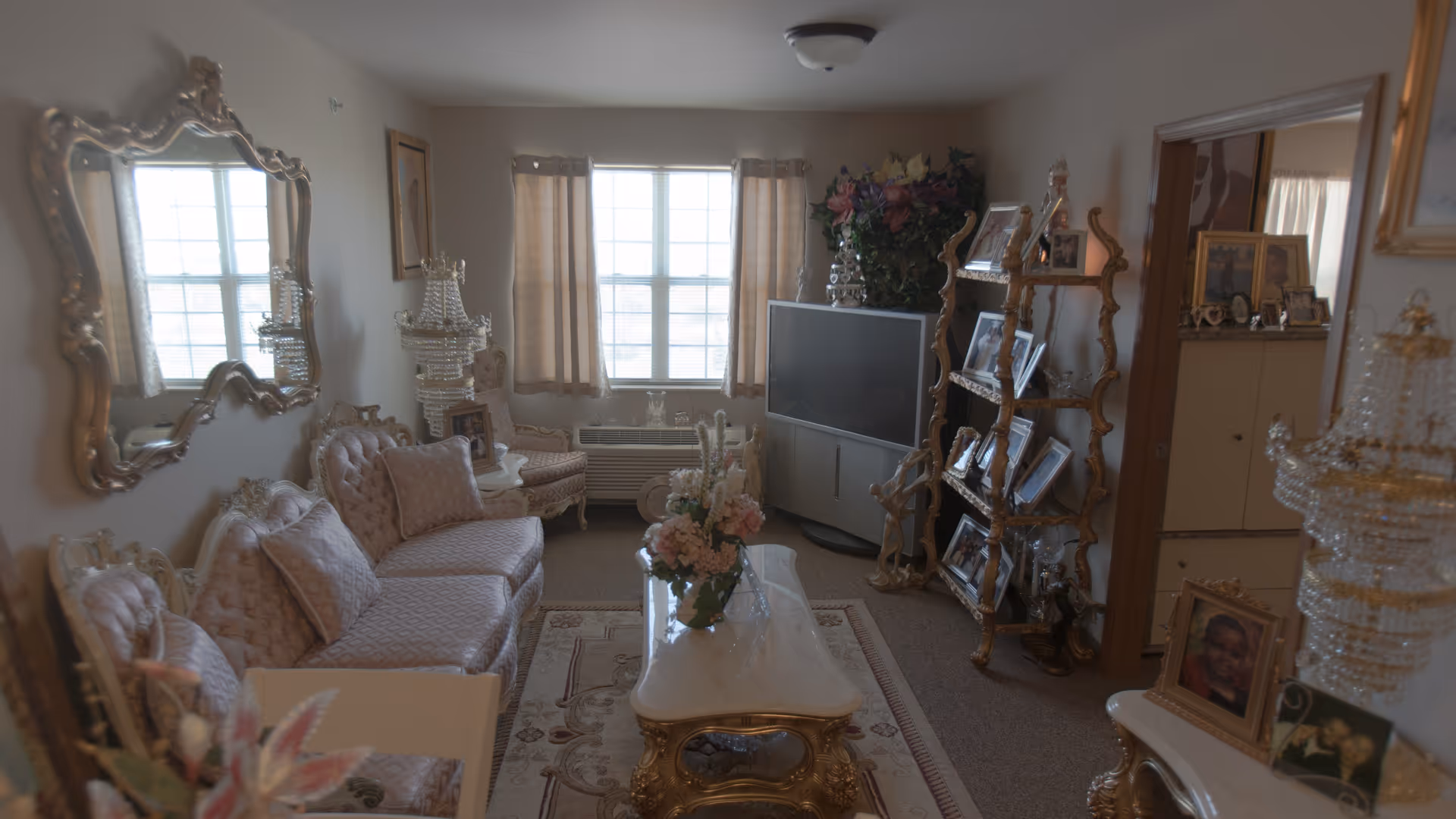A living room with ornate, vintage-style furniture including a tufted sofa and chairs with pink upholstery. A large decorative mirror hangs on the left wall above the sofa. In the center is a white coffee table with gold accents and a floral arrangement. A tall, gold-framed shelving unit on the right holds multiple framed photos. A flat-screen TV is positioned near a window with beige curtains. The room has a carpeted floor and soft lighting.