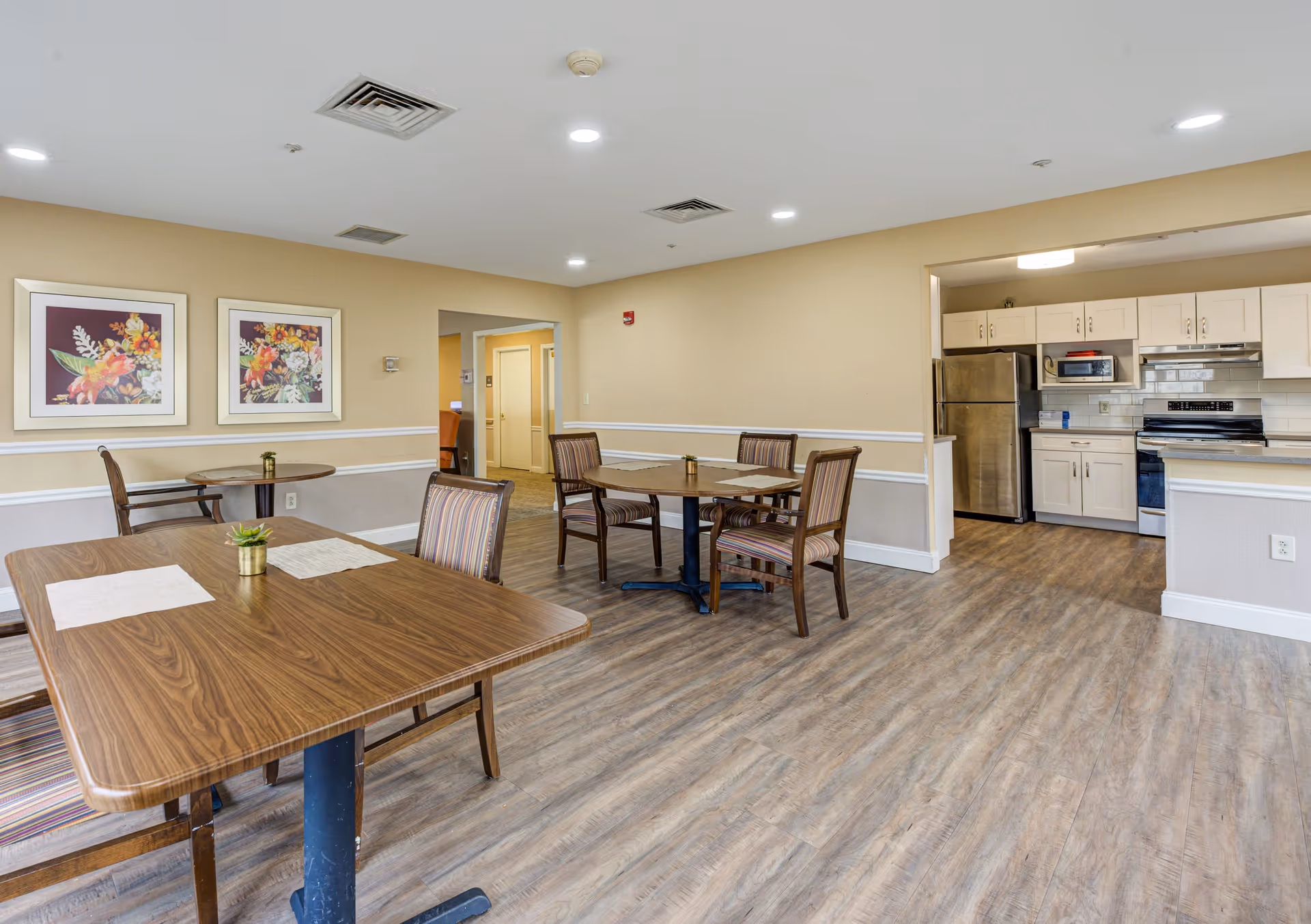A dining area in a senior living facility with wooden tables and striped cushioned chairs. The room has beige walls with white trim, two framed floral paintings on the wall, and a kitchen area with white cabinets and stainless steel appliances in the background. The floor is wood-patterned, and the ceiling has recessed lighting.