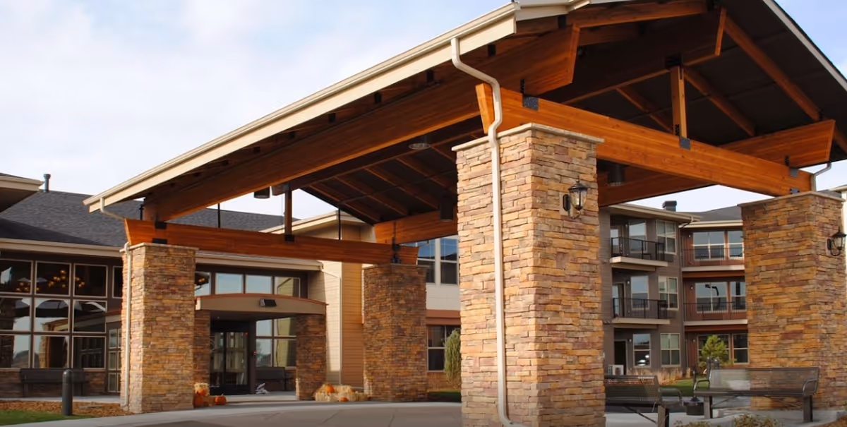 Entrance of a senior living facility with a large covered driveway supported by stone pillars and wooden beams. The building has multiple floors with balconies and large windows. There are benches and some landscaping visible near the entrance.