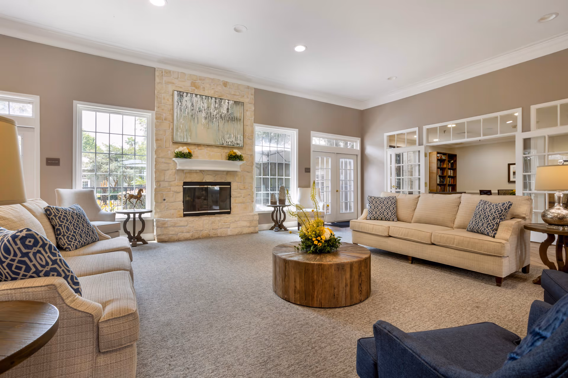 A spacious and well-lit senior living facility common area with beige sofas adorned with blue patterned pillows, a round wooden coffee table with a floral arrangement, a stone fireplace with a painting above it, large windows letting in natural light, and a glass door leading outside.