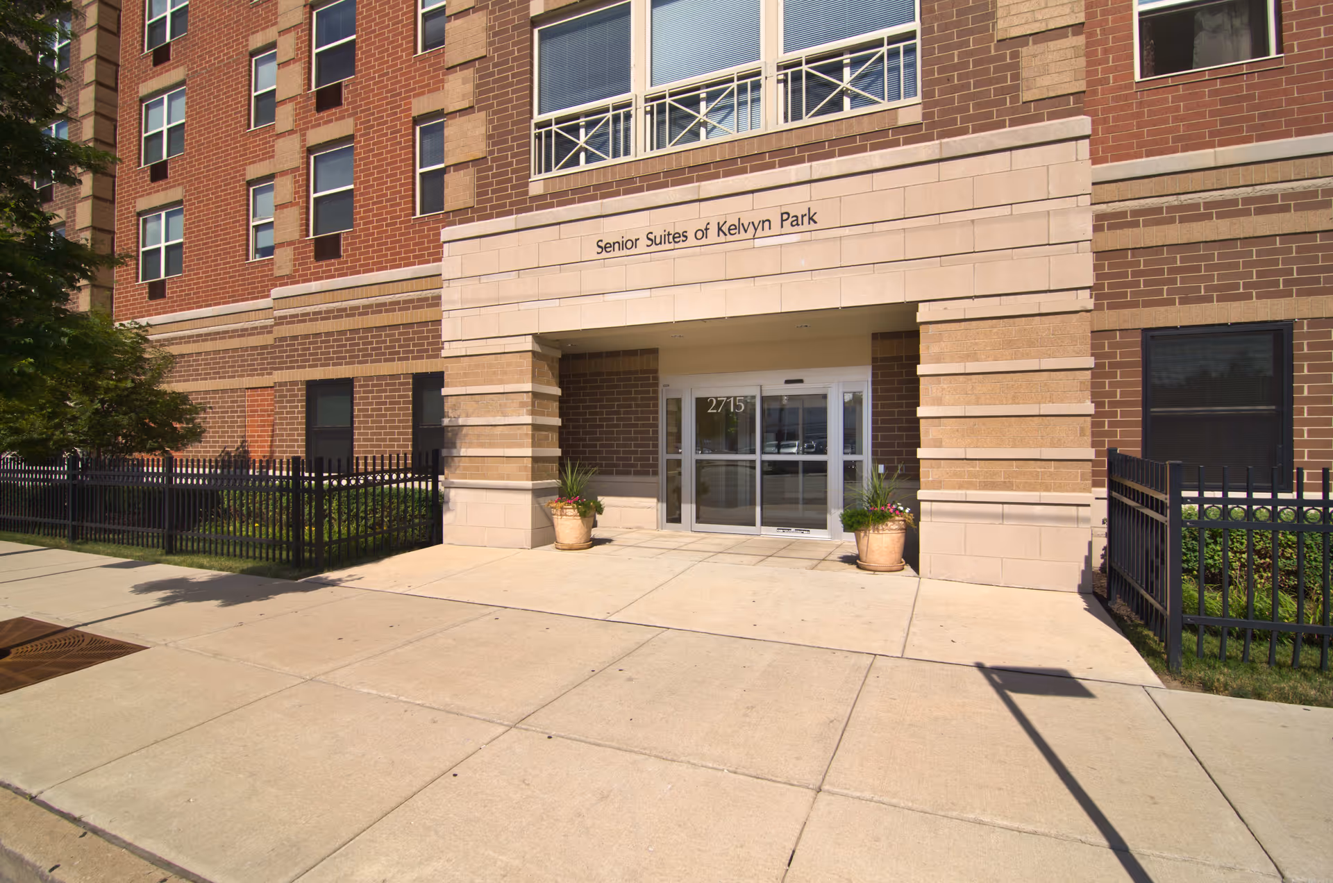 Exterior front entrance of a multi-story brick building with the sign 'Senior Suites of Kelvyn Park' above the doorway. The entrance has glass double doors with the number 2715 above them, flanked by two large potted plants. There is a sidewalk and black metal fencing on either side of the entrance.