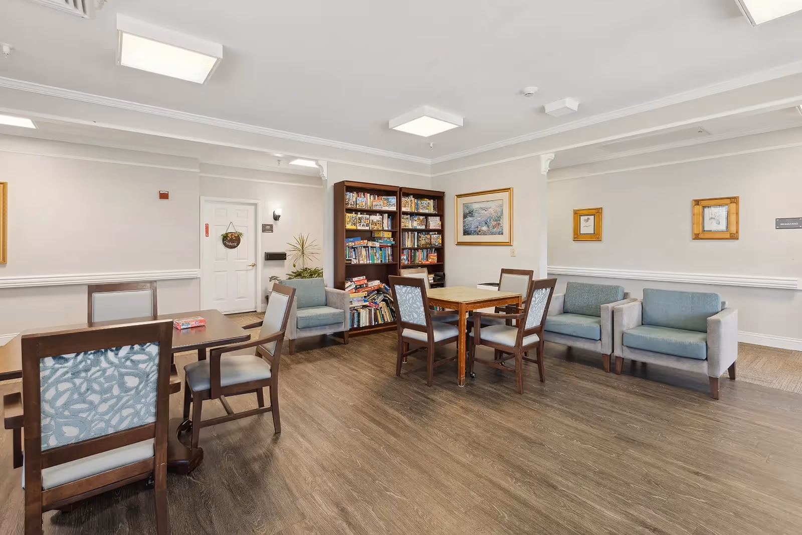 A well-lit common area in a senior living facility featuring wooden tables and chairs, a bookshelf filled with board games and books, two armchairs, and framed artwork on the walls. The room has a wood floor and white walls with crown molding.