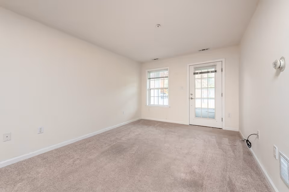 Empty room with beige carpet, white walls, a window, and a glass door leading outside. The room has electrical outlets and a thermostat on the wall.
