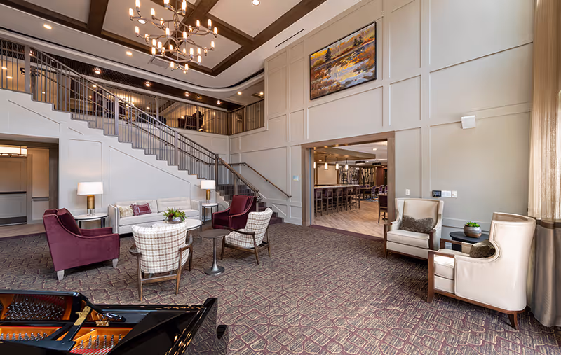 Spacious lobby lounge with grouped seating, a grand staircase, chandelier, and a piano in the foreground.