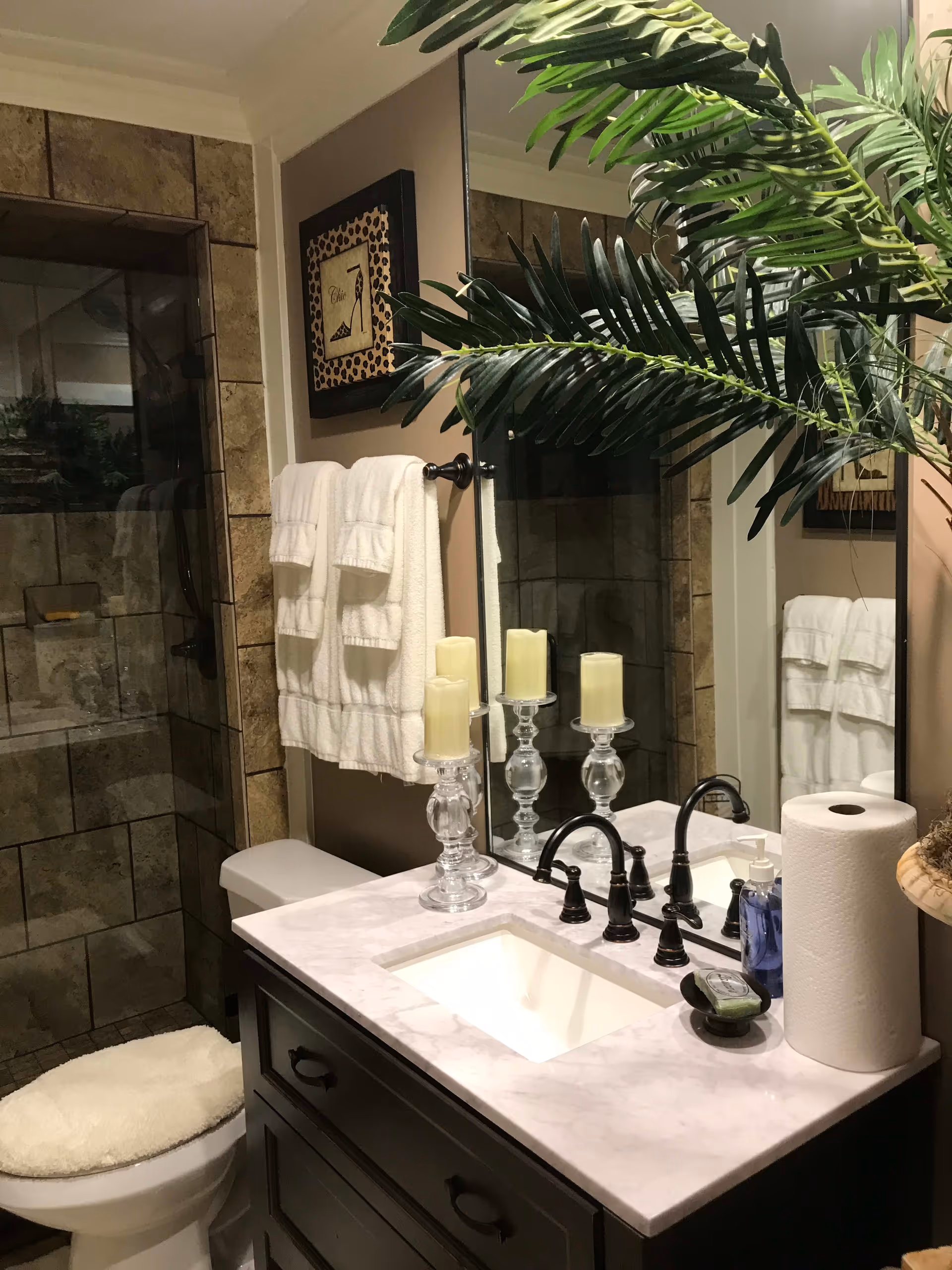 A bathroom with a marble countertop vanity featuring a sink, black faucet, three cream-colored candles on glass holders, a soap dispenser, and a roll of paper towels. There is a large mirror above the sink, a toilet with a cushioned seat cover, a towel rack with white towels, a framed picture on the wall, and a glass-enclosed shower with tiled walls. A large green plant is partially visible on the right side.