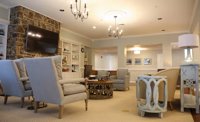 A cozy living room area in an assisted living facility featuring several upholstered armchairs arranged around a round wooden coffee table. A flat-screen TV is mounted on a stone fireplace wall. The room is softly lit by ceiling chandeliers and recessed lighting, with neutral-colored walls and carpet. Decorative shelves and framed pictures adorn the walls.