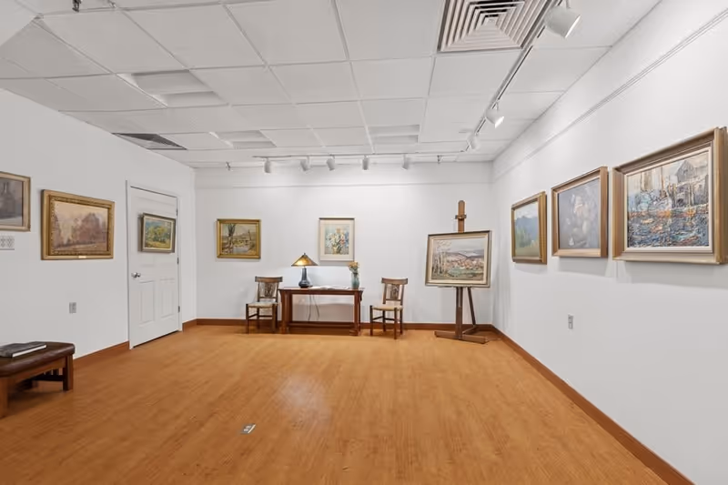 An art gallery room with wooden flooring and white walls displaying framed paintings. There is a small wooden table with a lamp and a vase with flowers, flanked by two wooden chairs. An easel holds a painting on the right side of the room. The ceiling has white tiles with track lighting illuminating the artwork.