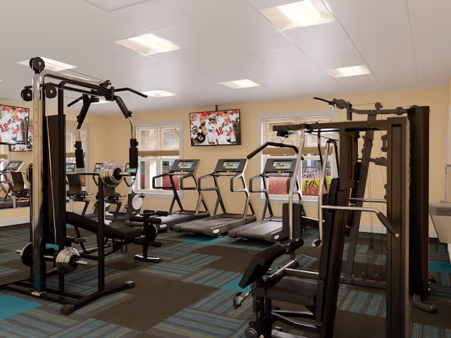 A fitness room with various exercise equipment including treadmills, weight machines, and a bench press. The room has large windows with blinds, a wall-mounted TV showing a football game, and a patterned carpet floor.