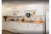 A clean and bright kitchen with white cabinets and drawers, a white microwave above a white stove, a dishwasher, a sink with a faucet, and a countertop decorated with a bowl of fruit, a vase of flowers, and a cookie jar. The walls are light-colored, and there is a paper towel dispenser mounted above the sink.