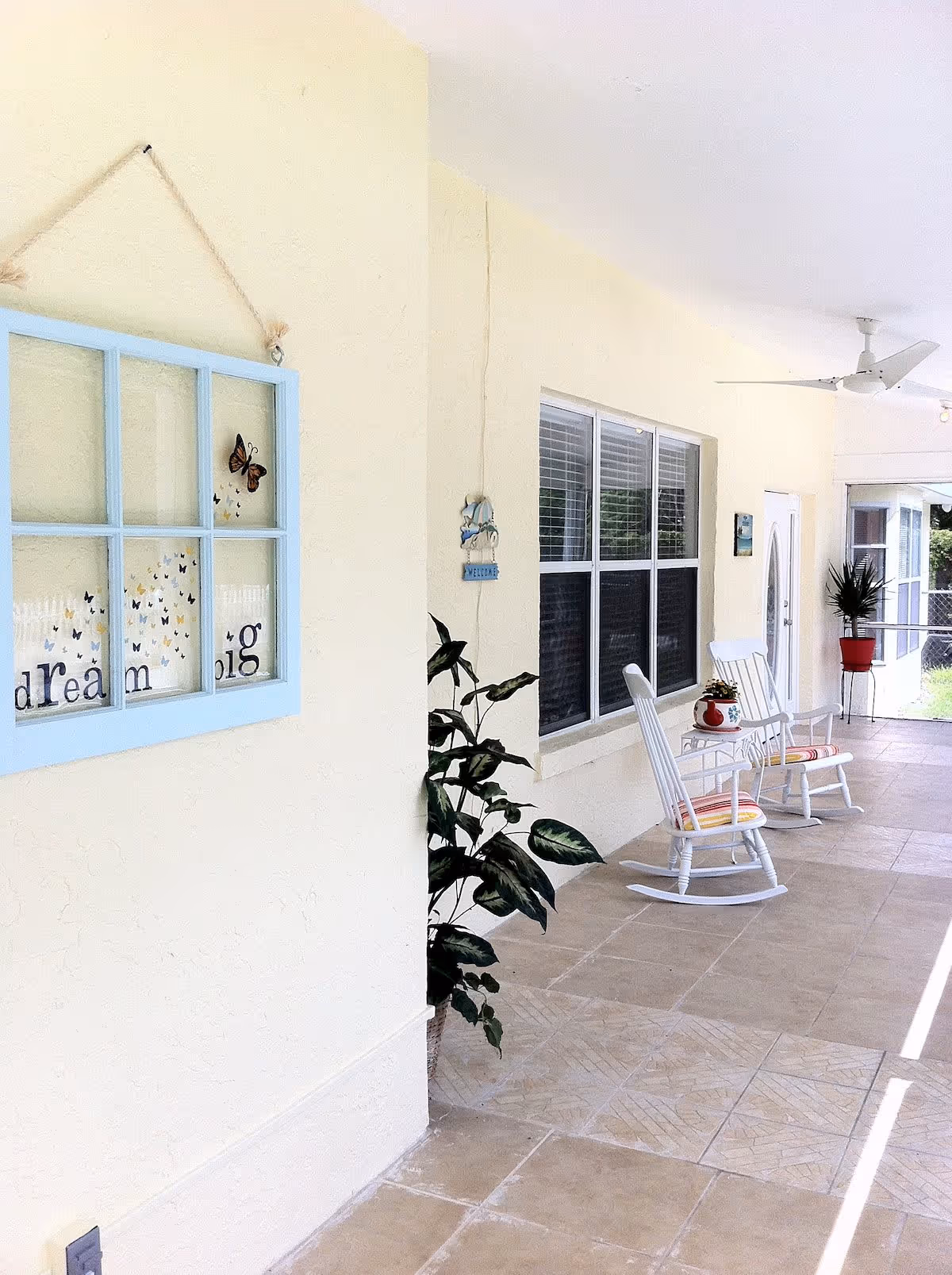 A bright and airy covered porch area with cream-colored walls and tiled floor. There are two white rocking chairs with striped cushions and a small white table between them holding a potted plant. A ceiling fan is mounted above. Decorative elements include a blue-framed window hanging with butterflies and the words 'dream big', a potted plant near the wall, and a welcome sign with dolphins. Large windows and a door lead into the building.