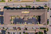 Aerial view of a large, rectangular senior living facility building with a central parking lot filled with cars. The building has a brown roof and is surrounded by roads and additional parking areas. The image shows the layout of the facility and its immediate surroundings.