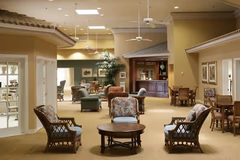 Spacious senior living common lounge with wicker armchairs around a coffee table and a small bar/reception area in the background.