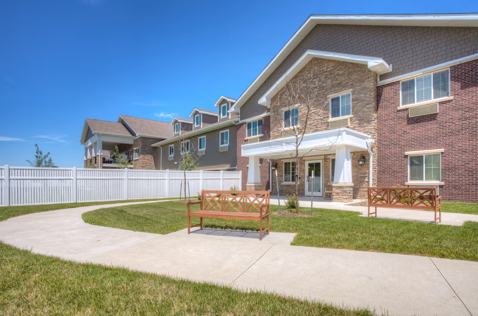 Exterior view of a senior living facility building with a stone and brick facade under a clear blue sky. There are two wooden benches on a curved concrete pathway surrounded by green grass and a white fence in the background.