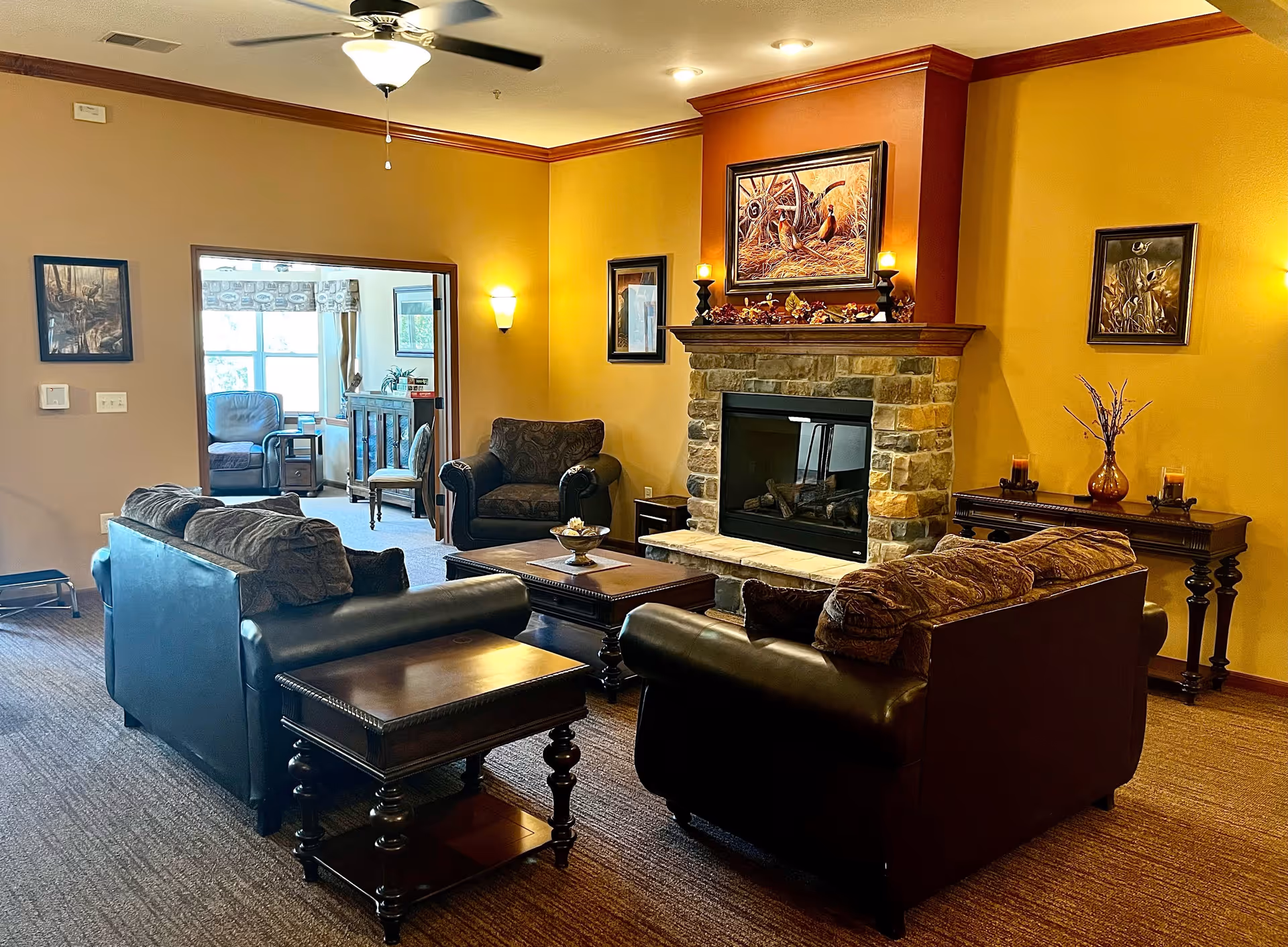 A cozy living room with two dark leather sofas and a matching armchair arranged around a wooden coffee table in front of a stone fireplace. The walls are painted warm yellow and decorated with framed artwork. A ceiling fan with a light hangs above, and a side table with candles and a vase is against the wall. An adjacent room with additional seating and large windows is visible through an open doorway.